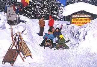 Eine Gruppe von Menschen mit Schlitten sitzt und steht an einem verschneiten Eingang, der mit einem gelben Schild "Beleuchtete Rodelbahn" gekennzeichnet ist, umgeben von Schnee und Bäumen.