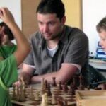 Ein Erwachsener und drei Kinder sitzen an einem Tisch und spielen Schach. Der Erwachsene schaut auf das Brett, während ein Kind eine Figur zieht und ein anderes Kind zusieht.