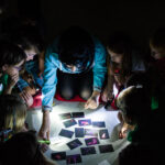 Eine Gruppe von Kindern und ein Erwachsener sehen sich in einem dunklen Raum mehrere Karten an, die von Taschenlampen beleuchtet werden.