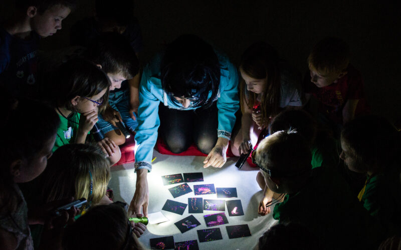 Eine Gruppe von Kindern und ein Erwachsener sehen sich in einem dunklen Raum mehrere Karten an, die von Taschenlampen beleuchtet werden.