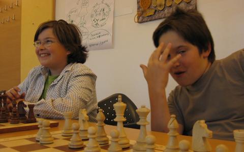 Zwei Kinder sitzen an einem Tisch und spielen Schach; eines lächelt, während das andere einen Teil seines Gesichts mit einer Hand verdeckt. Die Schachfiguren sind auf dem Brett vor ihnen angeordnet.