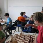 Kinder und Erwachsene spielen Schach an Tischen in einem Klassenzimmer, wobei ein Erwachsener den Raum beobachtet.