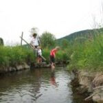 Zwei Kinder und ein Erwachsener stehen an einem kleinen Bach, die Kinder waten im Wasser, und auf beiden Seiten gibt es grasbewachsene Ufer.