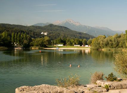Ein ruhiger See mit zwei Schwimmern im Wasser, umgeben von Bäumen und Bergen unter einem klaren Himmel. Am Ufer sind Gebäude und in der Ferne ein Schloss zu sehen.