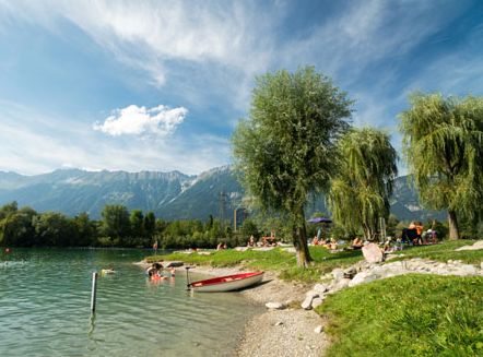 Menschen entspannen sich an einem Seeufer mit einem kleinen Boot auf dem Kiesstrand, Bäumen als Schattenspender und Bergen im Hintergrund unter einem teilweise bewölkten Himmel.