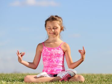 Ein junges Mädchen in einem rosa Oberteil sitzt im Schneidersitz im Gras, die Augen geschlossen, und meditiert mit den Händen in einer Mudra-Geste unter einem klaren blauen Himmel.