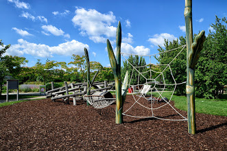 Spielplatz im Freien mit Klettergerüsten aus Holz, einem Seilnetz und grünen Bäumen unter einem blauen Himmel mit vereinzelten Wolken.