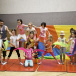 Eine Gruppe von Kindern und ein Erwachsener posieren auf Turnmatten in einer Halle, einige halten bunte Tücher in der Hand, und im Hintergrund ist ein Regenbogen zu sehen.
