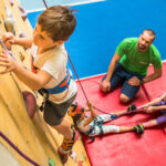 Ein kleiner Junge klettert an einer Indoor-Felswand, während ein Mann die Aufsicht führt und zwei Kinder auf Matten mit Kletterseilen umgehen.