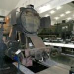 Eine verrostete, ausgemusterte Dampflokomotive steht in einem Innenraum auf einem Gleisabschnitt, während im Hintergrund ein anderer Zug zu sehen ist.