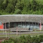 Ein großer, halbkreisförmiger Ringlokschuppen mit mehreren Türen, umgeben von Gleisen und Grünanlagen, vor dem eine rote Lokomotive zu sehen ist.