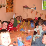 Eine Gruppe von Kindern sitzt bei einer Party um einen geschmückten Tisch mit Getränken und Luftballons; im Hintergrund sind Papierhüte und ein Fernseher zu sehen.