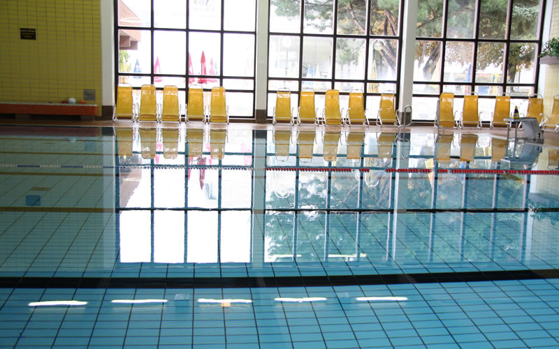 Ein Hallenbad mit klarem Wasser, blauen Kacheln und einer Reihe gelber Stühle vor großen Fenstern, die das Tageslicht hereinlassen.
