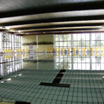 Hallenbad mit ruhigem Wasser, gefliestem Boden, großen Fenstern und Reihen von gelben Stühlen an den Wänden.