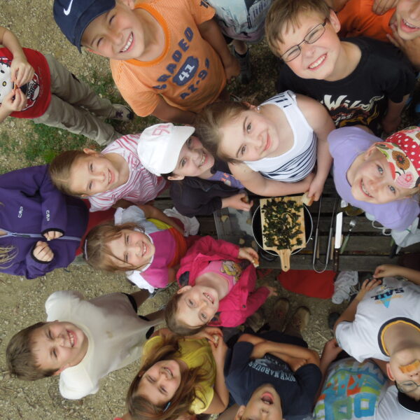 Eine Gruppe von Kindern steht im Freien in einem Kreis und blickt in die Kamera, in der Mitte steht eine Schale mit Essen auf einem Holztisch.
