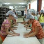 Eine Gruppe von Personen mit Haarnetzen arbeitet an einem langen Tisch in einer Großküche oder Bäckerei.