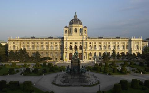 Ein großes, verziertes Gebäude mit einer zentralen Kuppel und gewölbten Fenstern, umgeben von einem Landschaftsgarten und einer Statue unter freiem Himmel.
