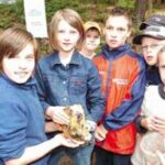 Eine Gruppe von Kindern im Freien, einige mit Jacken, steht dicht beieinander, während ein Kind einen großen Stein oder ein Mineral in der Hand hält.