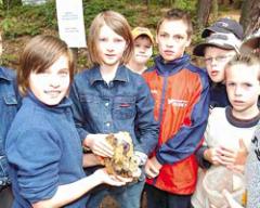 Eine Gruppe von Kindern im Freien, einige mit Jacken, steht dicht beieinander, während ein Kind einen großen Stein oder ein Mineral in der Hand hält.