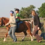 Ein Kind reitet auf einem Pony, wobei ein Erwachsener das Pony führt und ein anderer daneben geht, während ein Kleinkind in einem eingezäunten Außenbereich hinterherläuft.