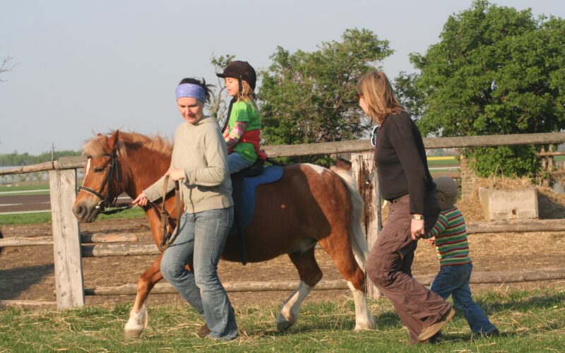 Ein Kind reitet auf einem Pony, wobei ein Erwachsener das Pony führt und ein anderer daneben geht, während ein Kleinkind in einem eingezäunten Außenbereich hinterherläuft.