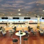 Eine moderne Bowlingbahn mit mehreren Bahnen und Overhead-Bildschirmen, mit Sitzbereichen mit Stühlen und Tischen im Vordergrund.