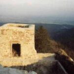 Ein kleines, verwittertes Steingebäude mit einer Tür steht an einem Berghang und überblickt eine ferne Landschaft unter einem dunstigen Himmel.