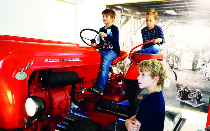 Drei Jungen interagieren mit einem alten roten Porsche Diesel Junior Traktor, der in einer Halle ausgestellt ist; zwei sitzen auf dem Traktor, während einer in der Nähe steht.