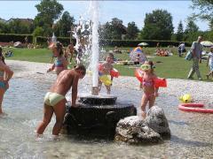 Kinder in Badeanzügen und mit Schwimmflügeln spielen in einem flachen Becken in einem Park um eine Wasserfontäne herum, während im Hintergrund Menschen auf dem Rasen faulenzen.