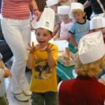 Eine Gruppe von Kindern mit Kochmützen aus Papier nimmt an einer Aktivität teil; ein Kind in einem gelben Hemd steht vorne mit erhobenen Händen und scheint aufgeregt zu sein.