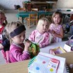 Fünf kleine Kinder sitzen um einen Tisch mit Snacks und Spielzeug in einer lockeren Innenumgebung mit Stühlen und Tischen im Hintergrund.