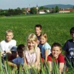 Eine Gruppe von acht Kindern sitzt und kniet auf einer grasbewachsenen Wiese mit einem Dorf und Hügeln im Hintergrund.