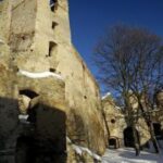 Eine hohe Steinruine steht im Sonnenlicht, teilweise mit Schnee bedeckt, mit blattlosen Bäumen und tiefblauem Himmel im Hintergrund.