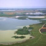 Luftaufnahme eines großen Sees, der von grünen Feldern, Ackerland und Vegetationsflecken unter einem dunstigen Himmel umgeben ist.