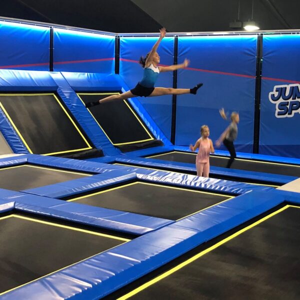 Fünf Kinder springen auf Trampolinen in einem Indoor-Trampolinpark namens "Jump Sport". Ein Kind führt einen Spagatsprung in der Luft aus.