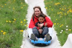 Ein Erwachsener und ein Kind fahren gemeinsam auf einem blauen Schlitten eine Betonrutsche hinunter, umgeben von Gras und gelben Blumen.