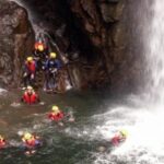 Eine Gruppe von Menschen mit Helmen und Schwimmwesten schwimmt und steht in einem Felsbecken unter einem Wasserfall.