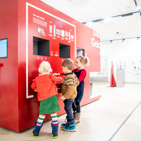 Drei kleine Kinder interagieren mit einem großen roten Touchscreen-Kiosk in einem hellen, modernen Ausstellungsraum.