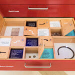 Eine offene Schublade mit der Aufschrift "Setz-Kasten" enthält unterteilte Fächer mit Münzen, Textkarten, Fotos und einem schwarzen Armband, das Teil eines Displays mit der Aufschrift "Geld-Puzzle" ist.