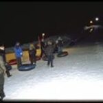 Eine Gruppe von Menschen steht nachts im Schnee, jeder mit einem aufblasbaren Schneeschlauch, in der Nähe eines mit Lichtern beleuchteten Tubing-Hügels.