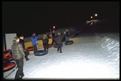 Eine Gruppe von Menschen steht nachts im Schnee, jeder mit einem aufblasbaren Schneeschlauch, in der Nähe eines mit Lichtern beleuchteten Tubing-Hügels.