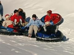 Mehrere Personen, darunter Kinder und Erwachsene, sitzen in Snowtubes und rutschen gemeinsam einen verschneiten Hügel hinunter.