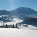 Ein schneebedecktes Tal mit verstreuten Bäumen und Häusern, umgeben von Bergen unter einem klaren blauen Himmel.