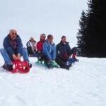 Fünf Personen sitzen auf Schlitten am Gipfel eines verschneiten Hügels und bereiten sich auf die Abfahrt mit dem Schlitten vor. Im Hintergrund sind Kiefern zu sehen.