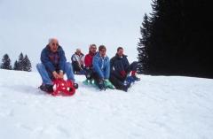 Fünf Personen sitzen auf Schlitten am Gipfel eines verschneiten Hügels und bereiten sich auf die Abfahrt mit dem Schlitten vor. Im Hintergrund sind Kiefern zu sehen.
