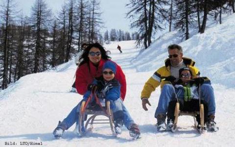 Eine vierköpfige Familie schlittelt einen verschneiten Hügel zwischen blattlosen Bäumen hinunter, alle tragen Winterkleidung und Sonnenbrillen.