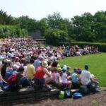 Eine große Gruppe von Kindern und Erwachsenen sitzt im Freien auf gestuften Sitzen und hört einer Person zu, die auf einer von Bäumen umgebenen Wiese steht und spricht.