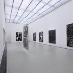 Eine moderne Kunstgalerie mit weißen Wänden zeigt mehrere abstrakte Schwarz-Weiß-Gemälde, darunter zwei hängende Leinwände in der Mitte des Raums.