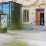 Eine Person betritt ein gelbes Gebäude mit gewölbten Fenstern und einer Glaskonstruktion im Außenbereich. In der Nähe des Eingangs ist ein buntes Plakat angebracht.