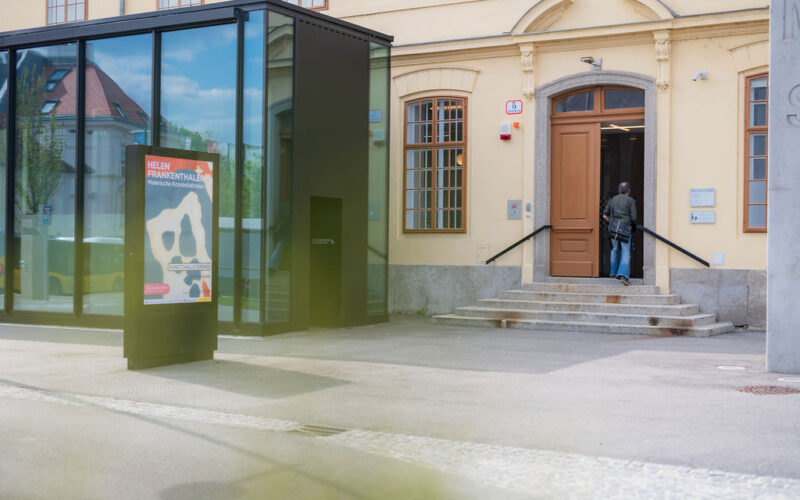 Eine Person betritt ein gelbes Gebäude mit gewölbten Fenstern und einer Glaskonstruktion im Außenbereich. In der Nähe des Eingangs ist ein buntes Plakat angebracht.